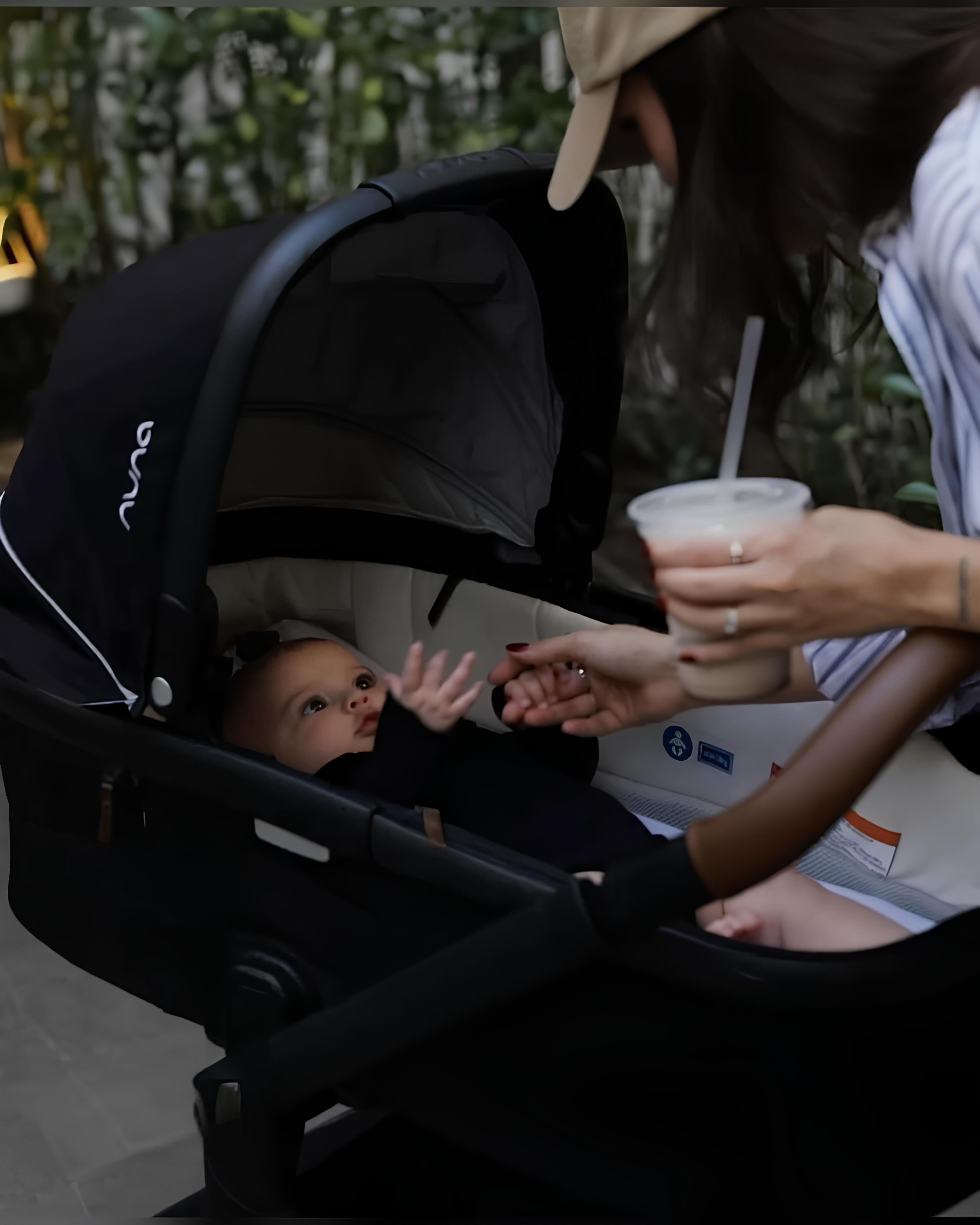 Person interacting with a baby in a stroller, holding a drink.